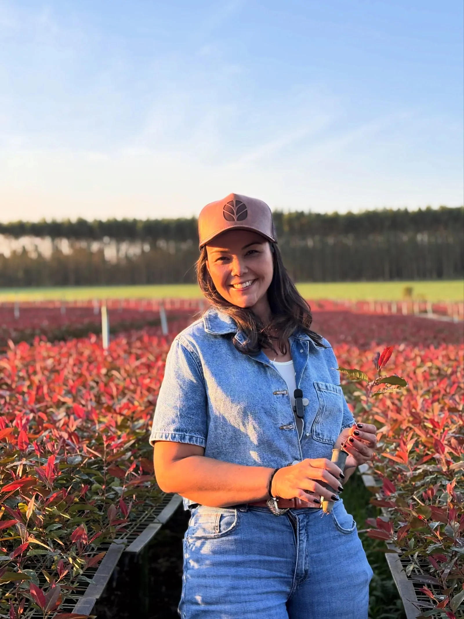 Bruna Satomi sorrindo no meio de um viveiro de mudas avermelhadas, ao fim da tarde